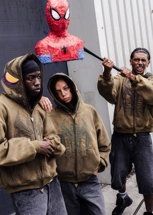 Three people wearing brown hoodies with green spider-themed graphics stand in front of a gray building wall. One person holds a large red Spider-Man piñata with a sword handle against their shoulder.